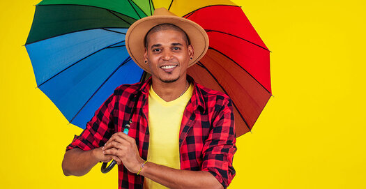 Mann mit dunklen kurzen Haaren, einem rot-schwarz karierten Hemd und einem bunten Regenbogen-Schirm in der Hand