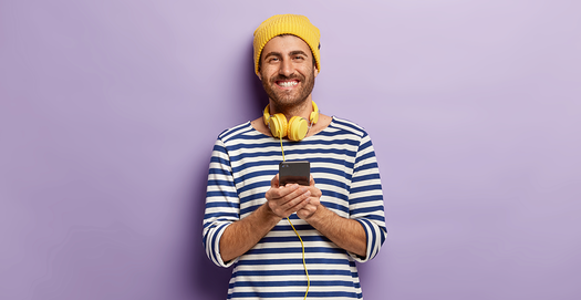 Ein junger Mann im Ringelshirt mit gelben Kopfhörern und gelber Mütze mit einem Handy in der Hand